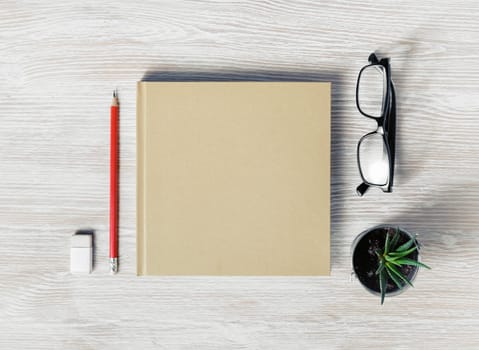 Photo of closed blank square booklet and stationery: glasses, pencil, eraser and plant on light wooden background. Template for placing your design. Flat lay.