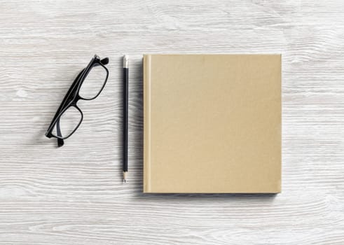 Photo of closed blank square book or notepad, glasses and pencil on wooden background. Template for placing your design. Flat lay.