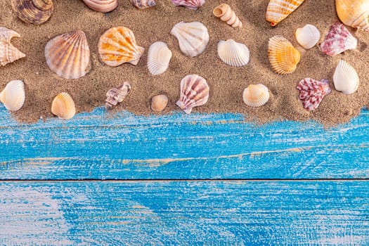 Summer time concept with sea shells on a blue wooden background and sand. Seashells frame on wooden background nautical border. Focus on seashells.
