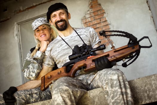 Smiling male and female military persons with crossbow weapon are sitting and looking at camera.