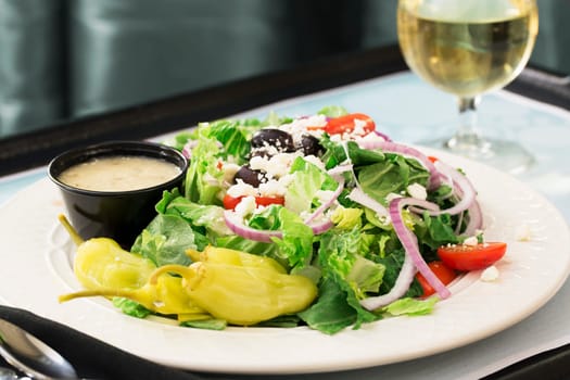 Fresh Greek salad on tray with dressing and glass of wine in the background.
