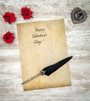 Antique preachment with Happy Valentines Day, 3 red roses, detailed silver quill stand in shape of hart accented by flat lying beautifully ornamented silver and black quill on rustic white painted oak