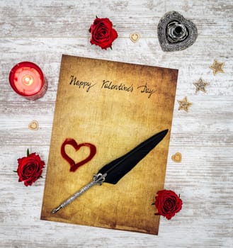 Antique preachment with written Happy Valentines Day, red candle, 3 red roses, red painted hart, wooden decorations, silver quill stand, ornamented silver quill on rustic white painted oak
