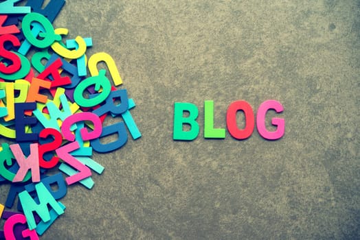 The colorful words "BLOG"" made with wooden letters next to a pile of other letters over grey background."