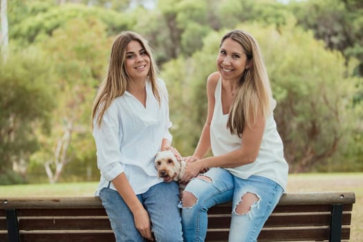 A mother and daughter are sitting by a park bench with their cute dog whilst smiling for a photo.