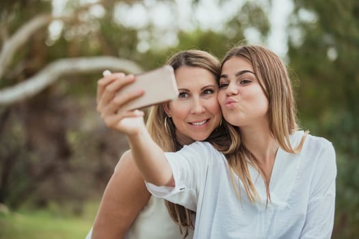 A mother and daughter are side by side in the park posing for a selfie with their dog.