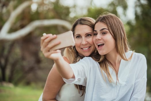 A mother and daughter are side by side in the park posing for a selfie with their dog.