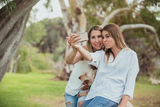 A mother and daughter are side by side in the park posing for a selfie with their dog.