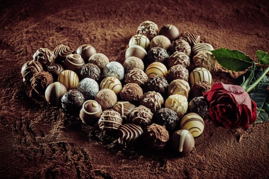 Heart shaped chocolate arrangement of luxury handmade pralines for Valentines Day with a single red rose for Love on a background of cocoa powder with vignette