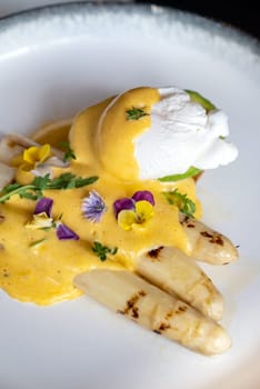 Gourmet grilled white asparagus with poached egg serve with hollandaise savory sauce decorate with edible colorful flowers