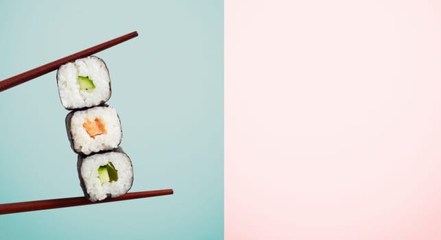 Chopsticks holding three seaweed wraps with rice and avocado over blue half of two tone background. Includes copy space.