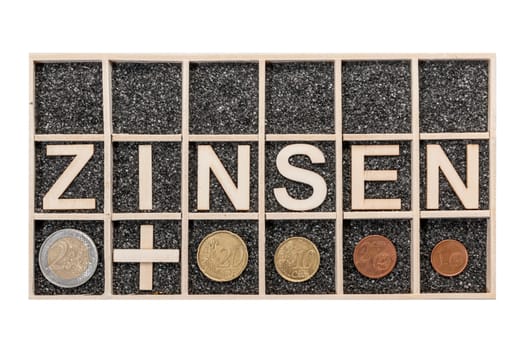 Plywood letters arranged in a collecting box with black decorative sand depicting the word ZINSEN and some Euro coins a nearby subjects in top view isolated on white.