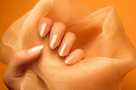 Female hand with glittered shiny bright golden colored nails is holding a beige textile on orange background, nail care and manicure concept.