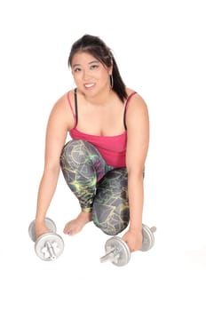 A beautiful young Chinese woman in exercising outfit kneeling on the.floor with two dumbbells, looking into camera, isolated for white .