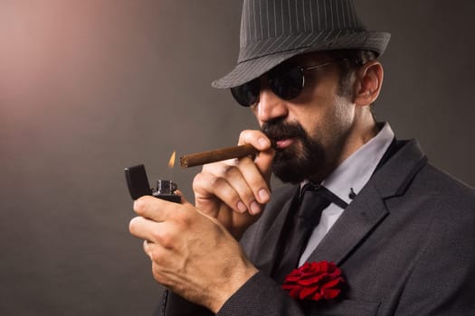 The elegant bearded gentleman in suit, hat and sunglasses is smoking cigar on gray background.