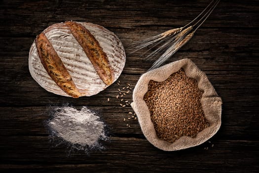 Still life with baking rye bread, ears, flour and bag of grains in a sackcloth on old wood background