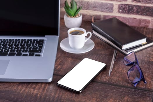 Laptop notebook with blank screen and cup of coffee and notepad pen and books and smartphone on wooden table. .Mock up on wooden table with laptop with blank screen, cup of coffee, pen, books and flower..Focus on smart-phone and glasses.