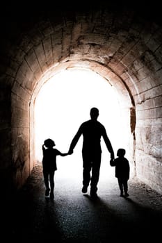 Father walking with his two little siblings children, boy and girl, towards the light at the end of a long dark tunnel