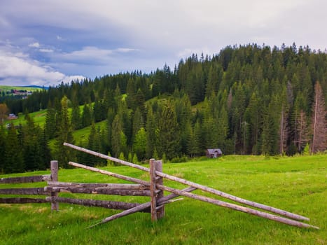 Picturesque spring mountains scene with wooden split rail fence across a green and lush pasture with a old house on the valley surrounded by coniferous woods. Yablunytsya village, Ukraine.