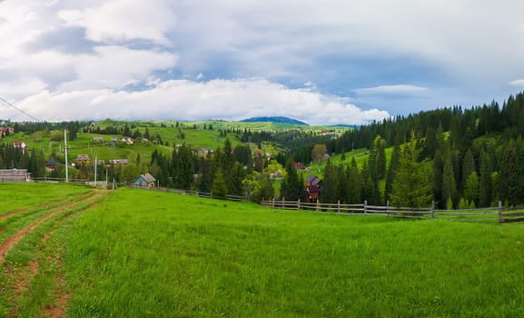 Picturesque spring mountains scene with wooden split rail fence across a green and lush pasture, a country road and old houses on the valley surrounded by pine woods. Yablunytsya, Ukraine.