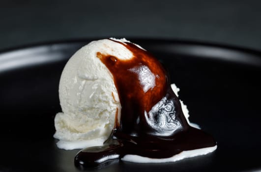 White cream ball on a black ceramic dish on a dark background, poured with chocolate sauce