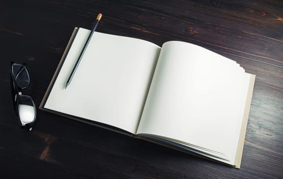 Blank booklet, pencil and glasses on dark wood table background.
