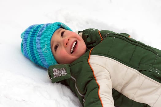 The little playful boy in warm clothing is lying down on the snow.