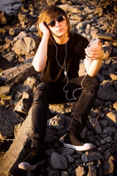 The trendy cool young guy with earphones is using phone, listening music and sitting on rocks outdoors.