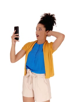 Beautiful young multi-racial woman standing in shorts holding her cell.phone in her hand is shocked about the message she got, isolated for.white background.