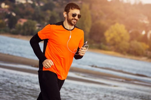 The smiling guy in orange sports clothing with earphones and sunglasses is jogging at the riverside, recreation concept.