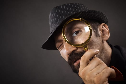 The old-fashioned detective with hat is looking through loupe or magnifier on dark gray background.