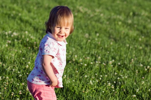 Smiling cute playful little girl is standing on green grass.
