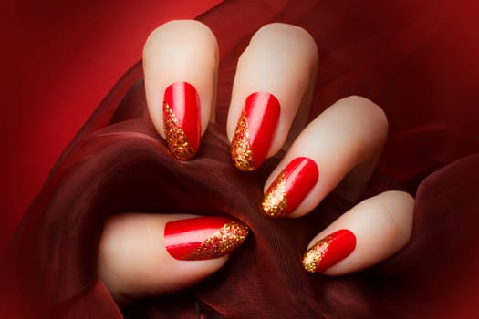 Female hand with beautiful red and golden glittered nails is holding a dark red textile on red background, manicure and nail care concept.