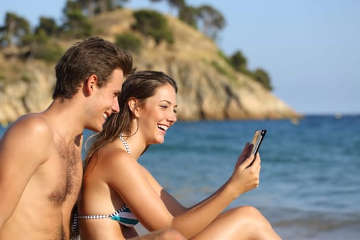 Side view portrait of a happy couple on the beach checking tablet on summer vacation