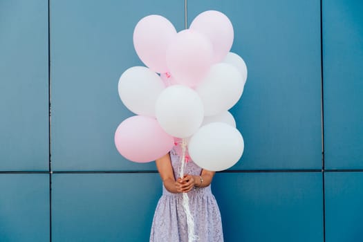 Young woman hiding her face with bunch of balloons, standing against blue wall. Dressed in tender dress. Outdoors.