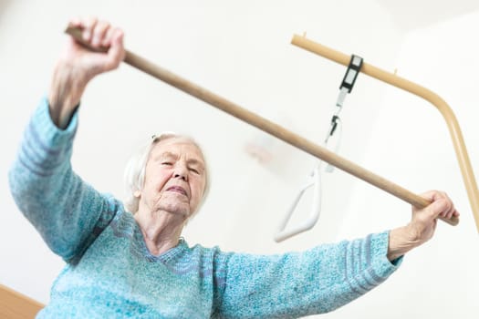 Elderly 96 years old woman exercising with a stick sitting on her bad. Geriatric health care home assisted support for older people concept. Care for the elderly.