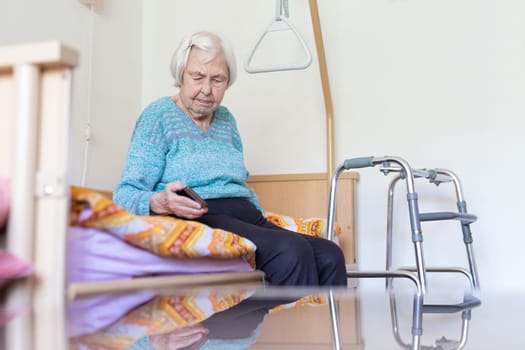Elderly 96 years old woman reading phone message while sitting on medical bed supporting her by holder. Senior woman using wireless internet connection on smart phone.