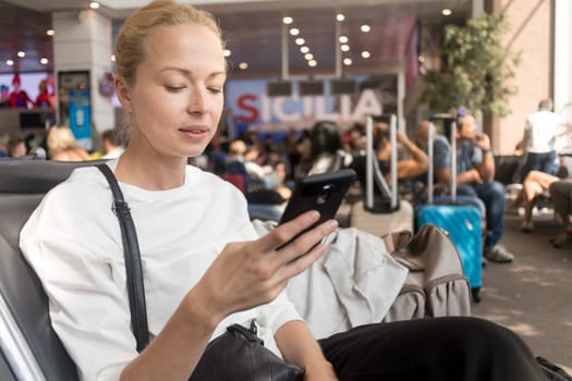 Casual blond young woman reading on her mobile phone while waiting to board a plane at the departure gates at the airport terminal. Travel concept.