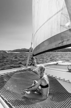 Beautiful woman relaxing on a summer sailing cruise, sitting and sunbathing in hammock of luxury catamaran sailing around Maddalena Archipelago, Sardinia, Italy. Black and white image.