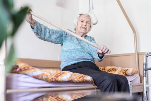 Elderly 96 years old woman exercising with a stick sitting on her bad. Geriatric health care home assisted support for older people concept. Care for the elderly.