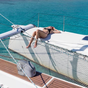 Woman in bikini tanning and relaxing on a summer sailin cruise, sitting on a luxury catamaran near picture perfect white sandy beach on Spargi island in Maddalena Archipelago, Sardinia, Italy.