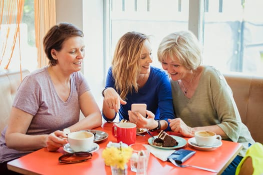 A group of three mature women having coffee and cake  they are looking at one of their phones.