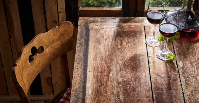 Old rustic wooden chair and table set with two glasses of red wine and a decanter in front of a window in a high angle view with copy space for placement of food