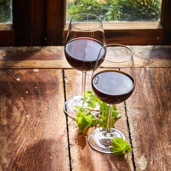 Two glasses of red wine with green vine leaves alongside a window on a rustic wooden worm ridden table in square format