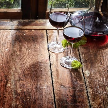 Two glasses of red wine with a decanter or carafe on a rustic wooden table alongside a window in a square high angle close up with copy space