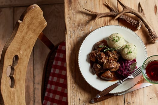 Serving of wild venison goulash with dumplings and red cabbage garnished with chopped fresh herbs on a rustic table set with deer antlers and wine