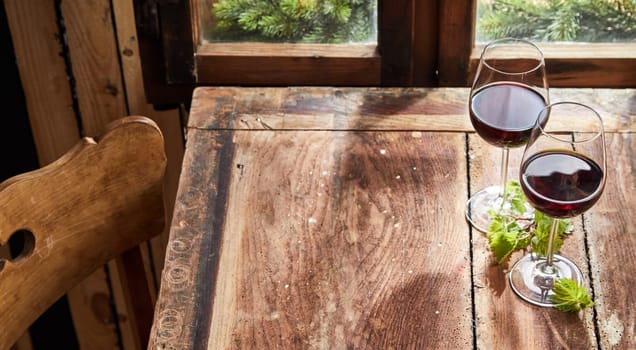 Two glasses of red wine on a rustic wooden table with fresh green leaves in front of a window and copy space for food placement in a high angle view
