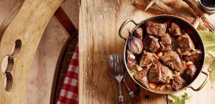 Delicious pot of wild venison goulash or ragout served on a rustic wooden table set with deer antlers and cutlery viewed top down in a panorama banner
