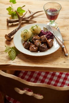 Plate of rich spicy wild venison goulash served with red cabbage and dumplings garnished with herbs on a rustic wooden table with red wine and deer antlers viewed over a chair