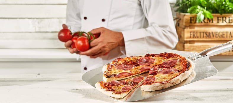 Chef with a fresh oven-fired Italian pepperoni pizza on a metal paddle standing in the background holding fresh tomatoes in his hands